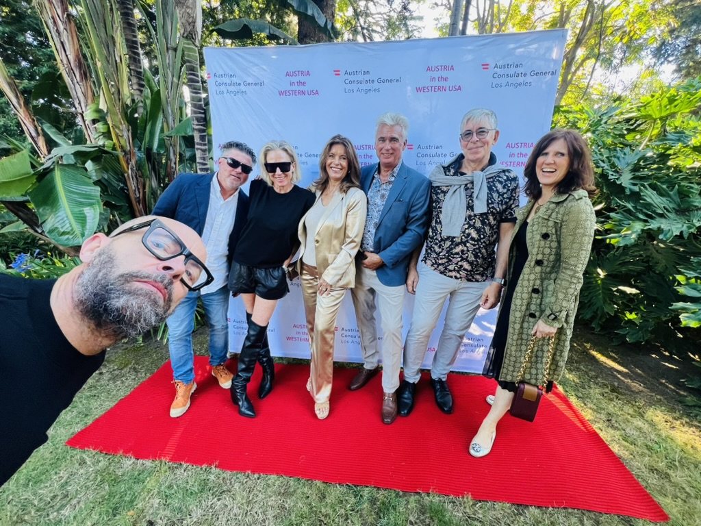 Claudia with a group of friends on a red carpet in a garden, in front of an Austrian Consulate General Los Angeles backdrop