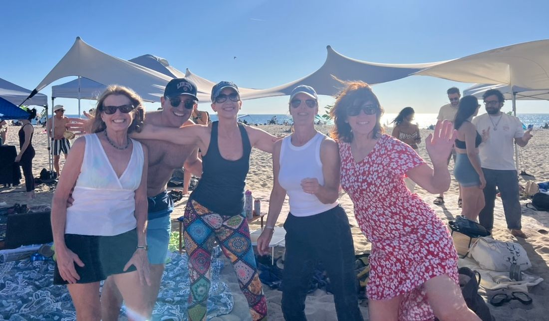 Friends laughing and waving under white shade canopies on a sunny beach, the ocean behind them
