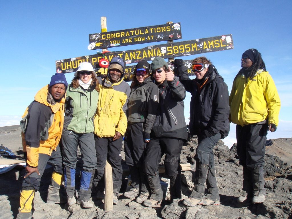 A group of climbers and guides in mountain jackets at the Uhuru Peak summit sign on Kilimanjaro, Tanzania, under a clear blue sky