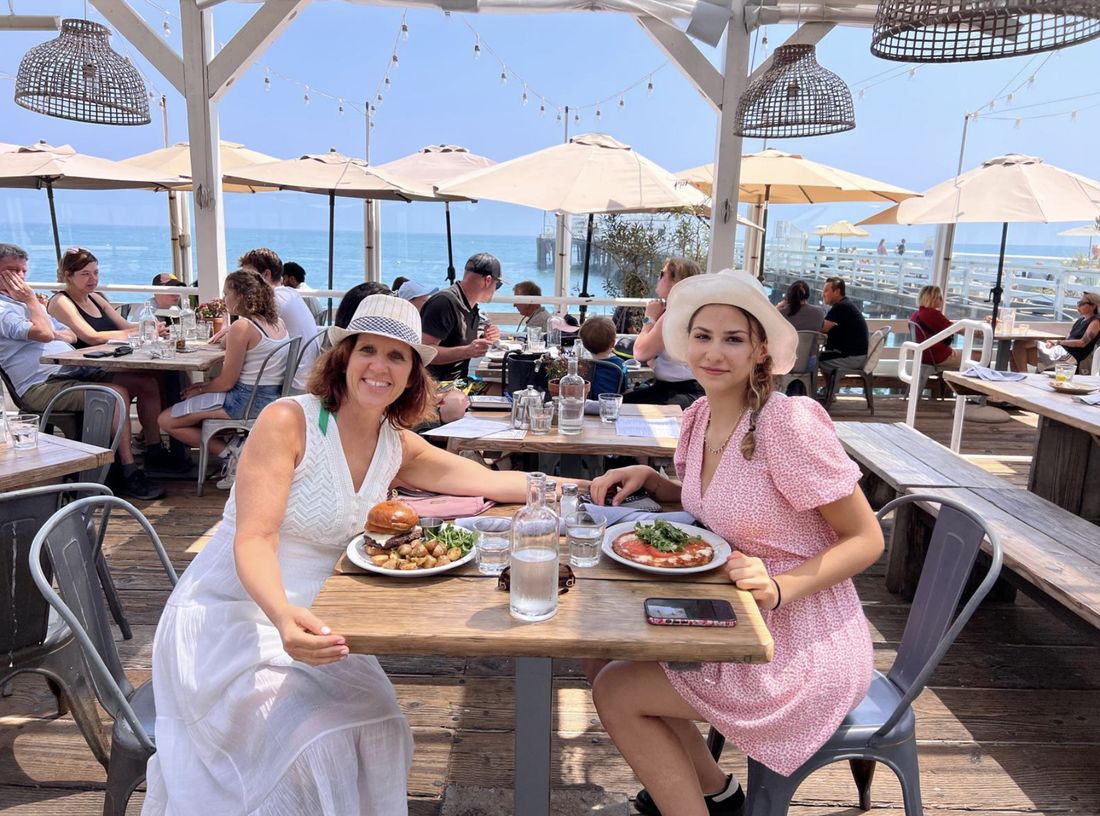 Claudia and her niece in sun hats at a table on a pier restaurant deck, the ocean behind them