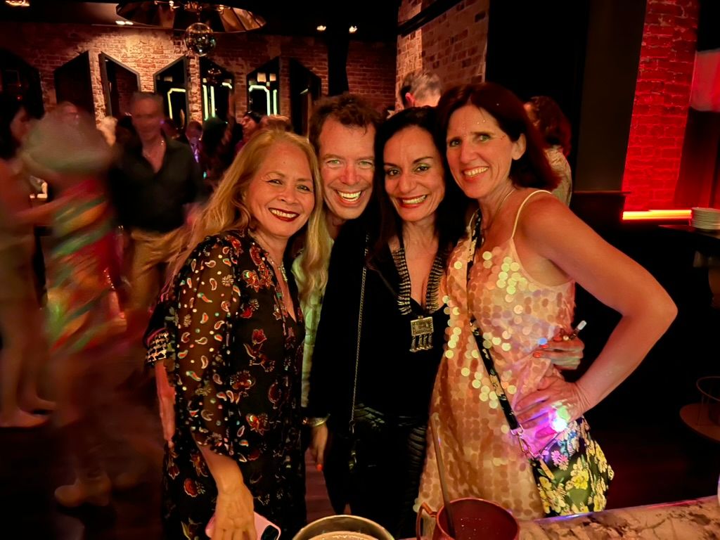 Claudia in a sequin dress with three friends, smiling close together in a busy club with brick walls