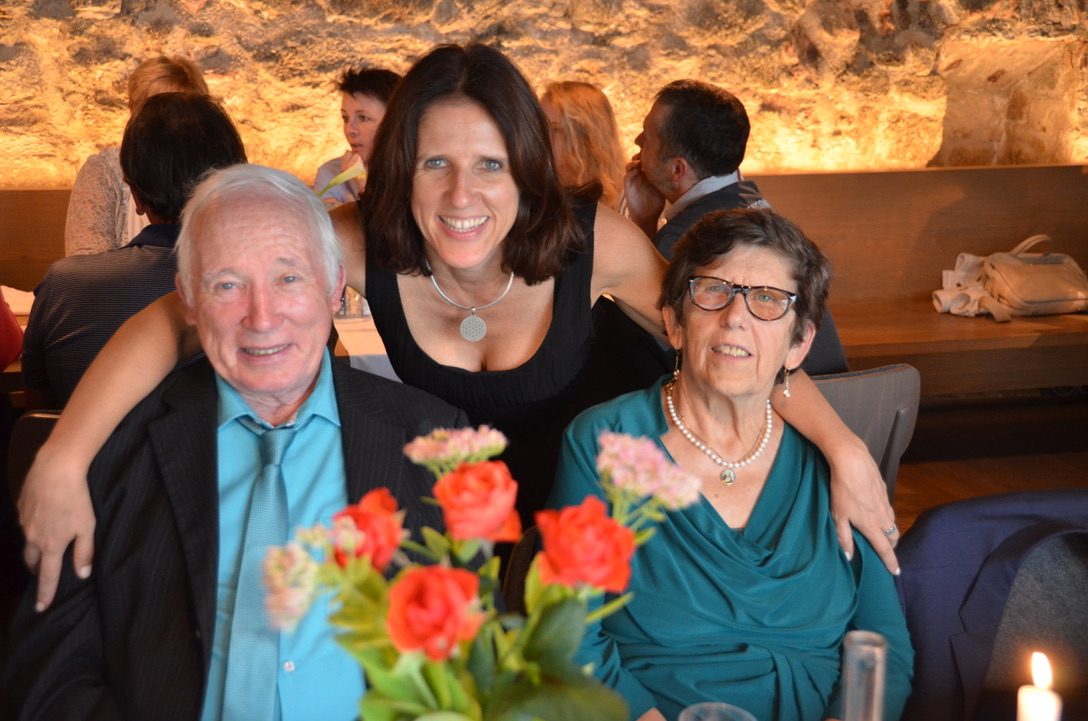 Claudia leaning in between her parents at a restaurant table, her father in a dark suit and her mother in teal, red roses in front of them