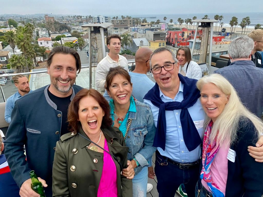 Claudia laughing with a group on a rooftop terrace, the beach and ocean behind them