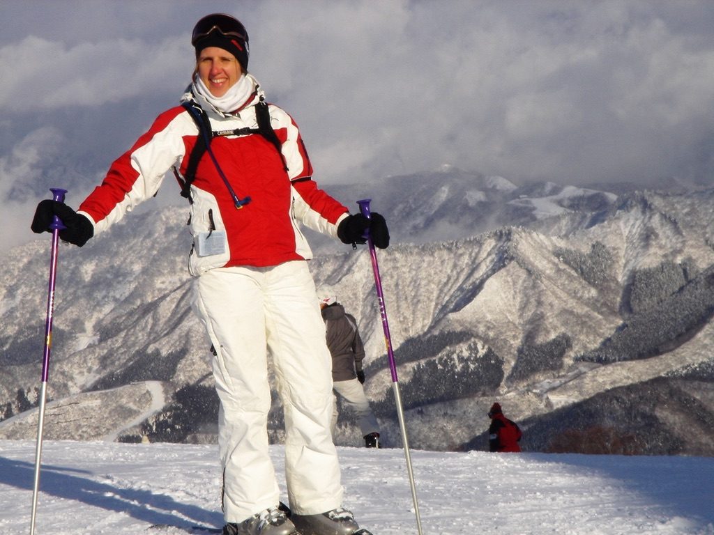 Claudia smiling on skis in a red and white jacket and white ski pants, poles out, snowy mountains and clouds behind her