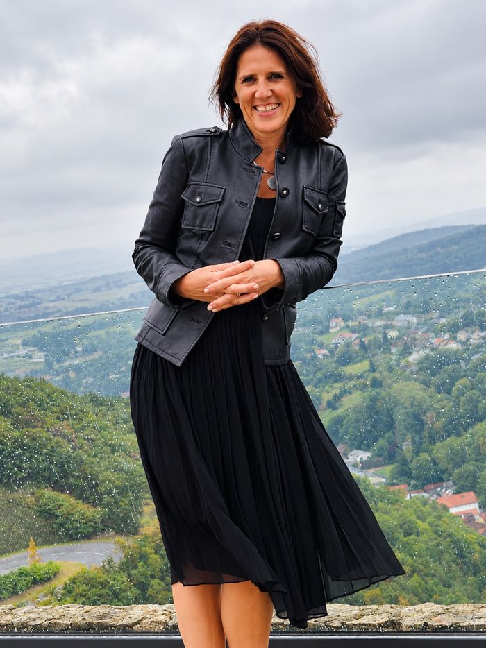 Claudia smiling on a terrace in a black leather jacket and pleated black dress, a green valley and village behind her